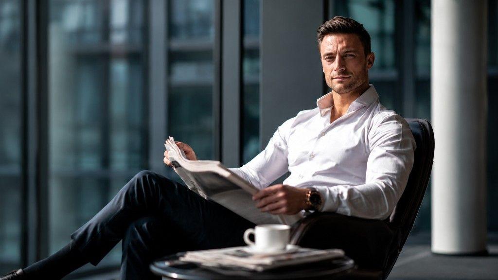 Man in a white open-collar shirt seated in a leather armchair in a bright office lounge, holding an open broadsheet newspaper, with coffee and a folded paper on a side table.